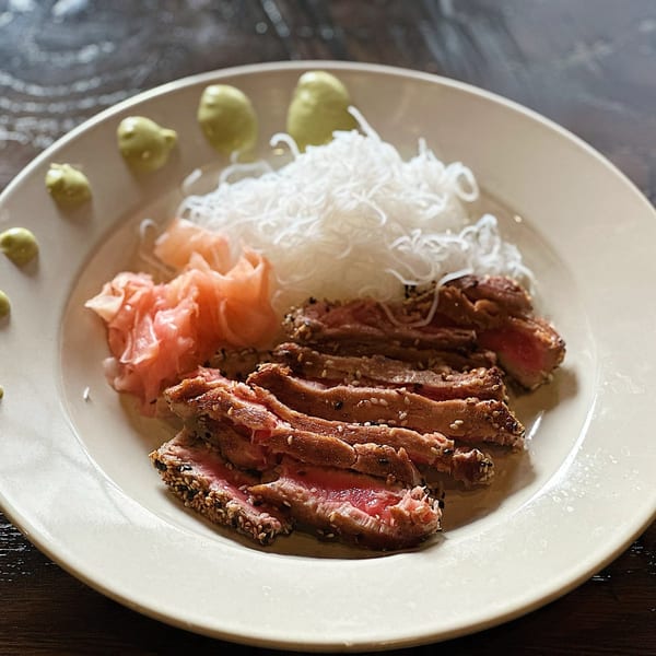 Seared tuna slices with sesame seeds, pickled ginger, rice noodles, and wasabi on a white plate.
