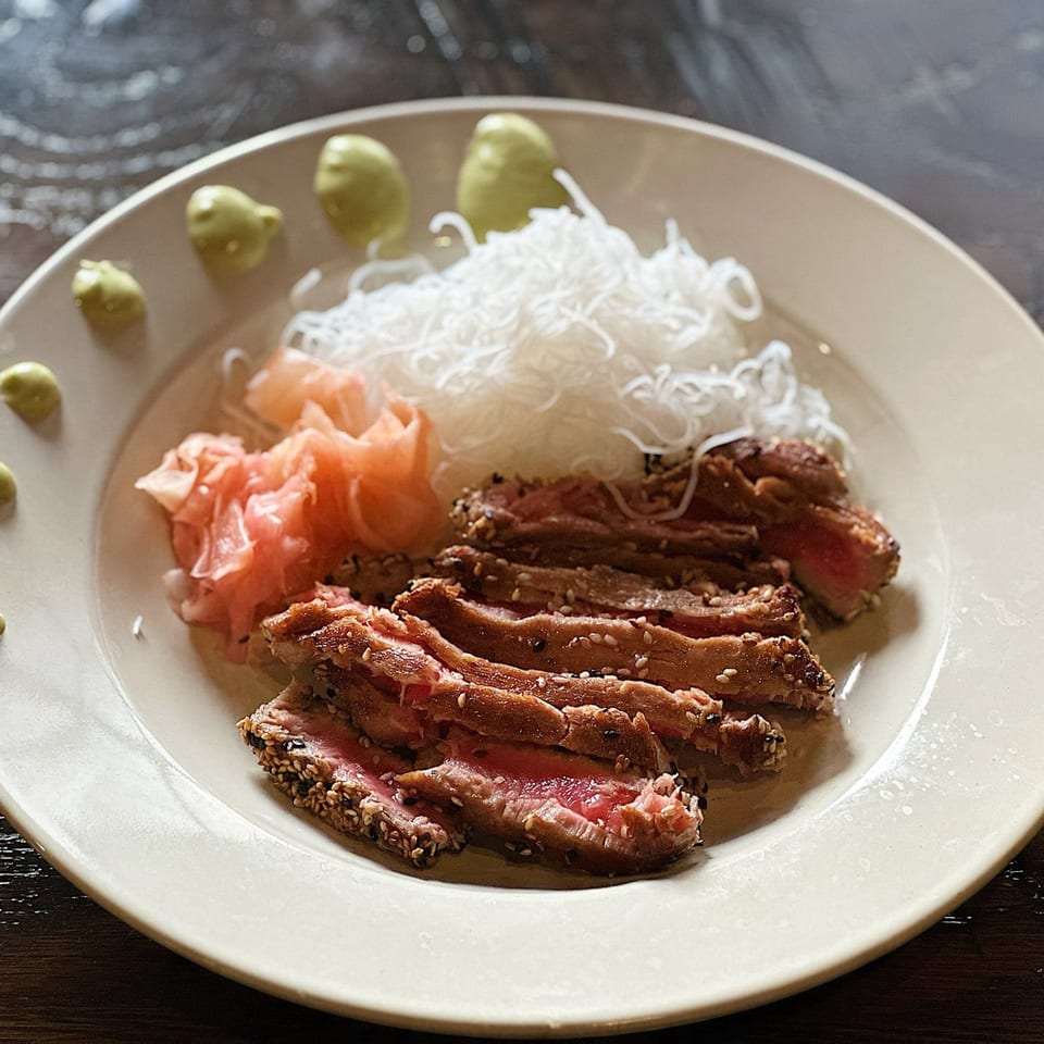 Seared tuna slices with sesame seeds, pickled ginger, rice noodles, and wasabi on a white plate.