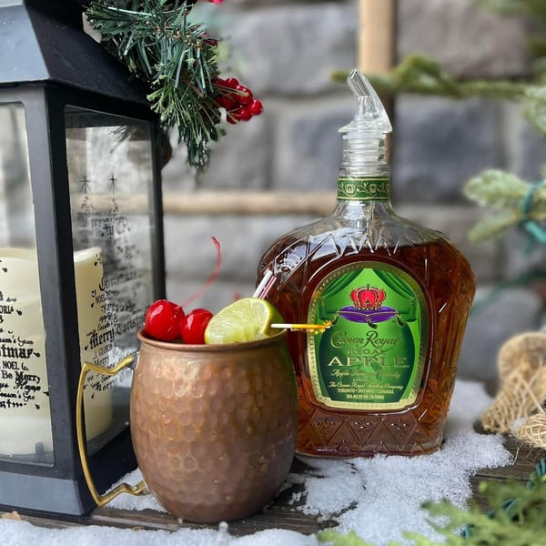 A copper mug cocktail with cherries and lime, next to a bottle of Crown Royal Apple on snow.