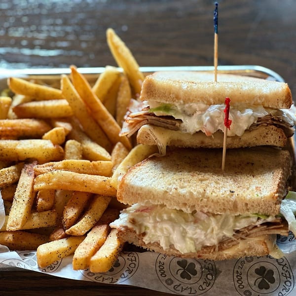 A club sandwich with melted cheese and coleslaw, served with seasoned fries on a metal tray.